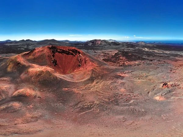 Paisaje volcánico árido con conos de lava rojo/naranja y terreno desolado