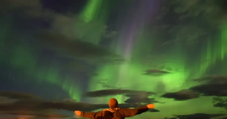Persona con brazos extendidos bajo aurora boreal de luz verde en cielo nocturno