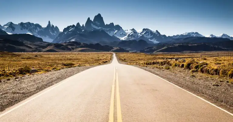 Ruta recta con línea amarilla central atravesando llanura hacia montañas nevadas