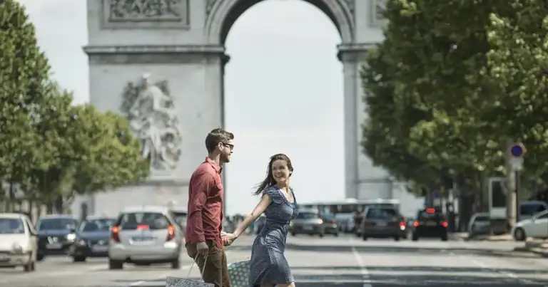Pareja caminando bajo arco de piedra blanca frente a avenida con árboles