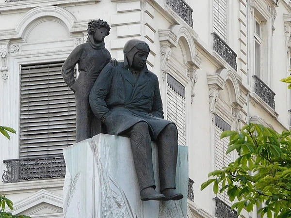 Estatua de bronce de hombre sentado en pedestal frente a edificio neoclásico