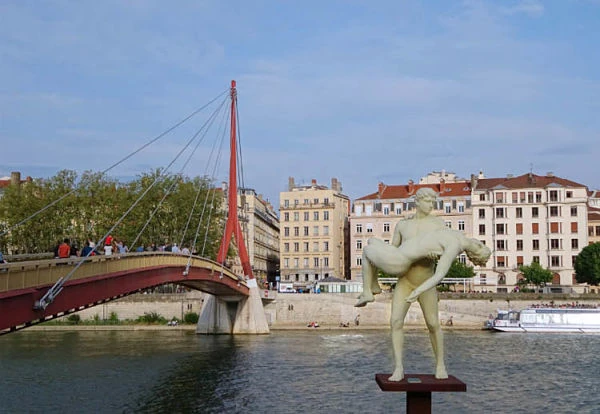 Puente rojo moderno con esculturas metálicas sobre río urbano y edificios históricos