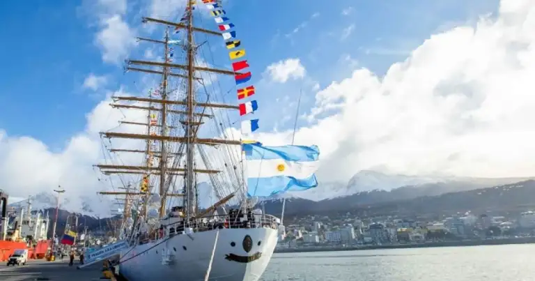 Velero histórico de velas en puerto con montañas y ciudad al fondo