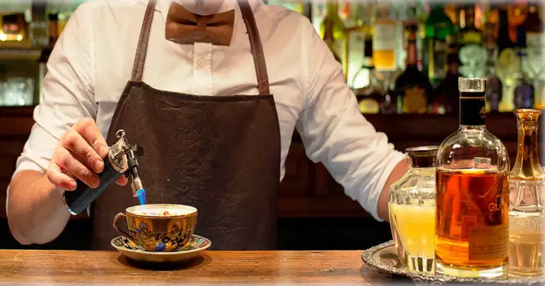 una persona preparando un coctel. Se ve una taza, una llamade fuego y una copa