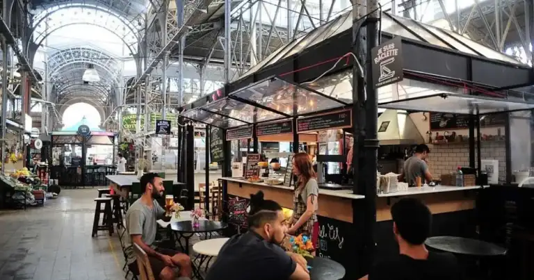 Mercado cubierto con estructura metálica, visitantes en bares y mesas