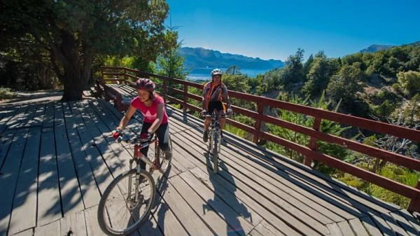 Dos ciclistas en pasarela de madera con montaña de fondo