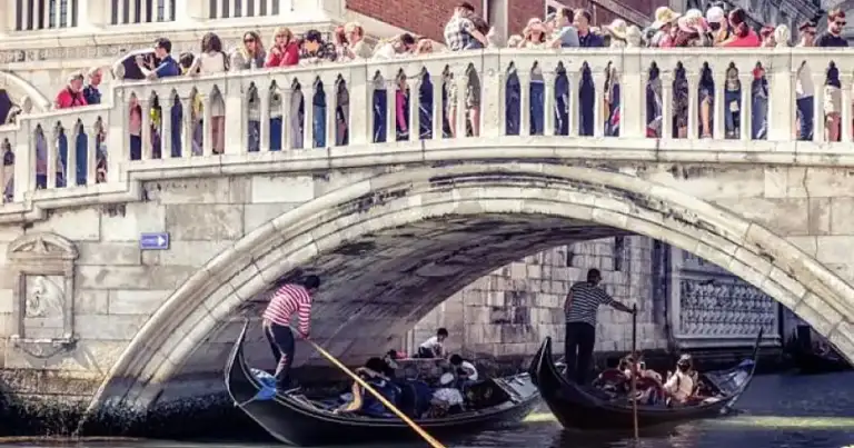 Gondolero en canal veneciano bajo puente arqueado con turistas en balcones