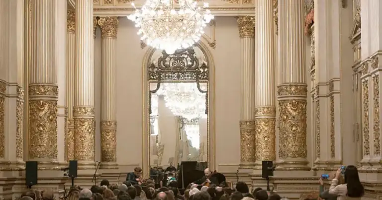 Salón ornamentado con columnas doradas, araña de cristal y público en concierto