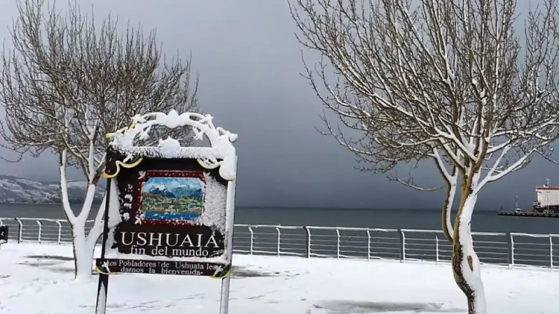 Cartel de Ushuaia con ramas nevadas y vista al agua al atardecer