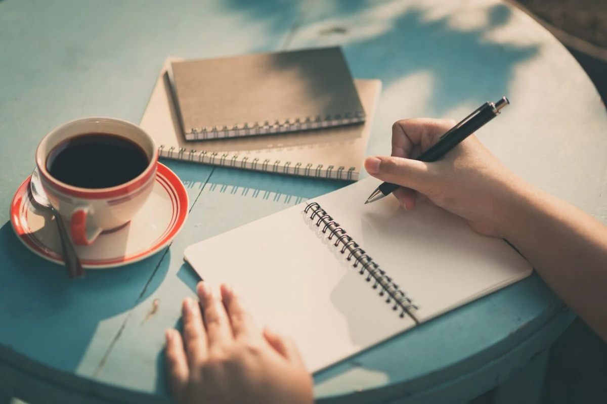 Persona escribe en cuaderno con taza de café, bolígrafo y cuadernos en mesa