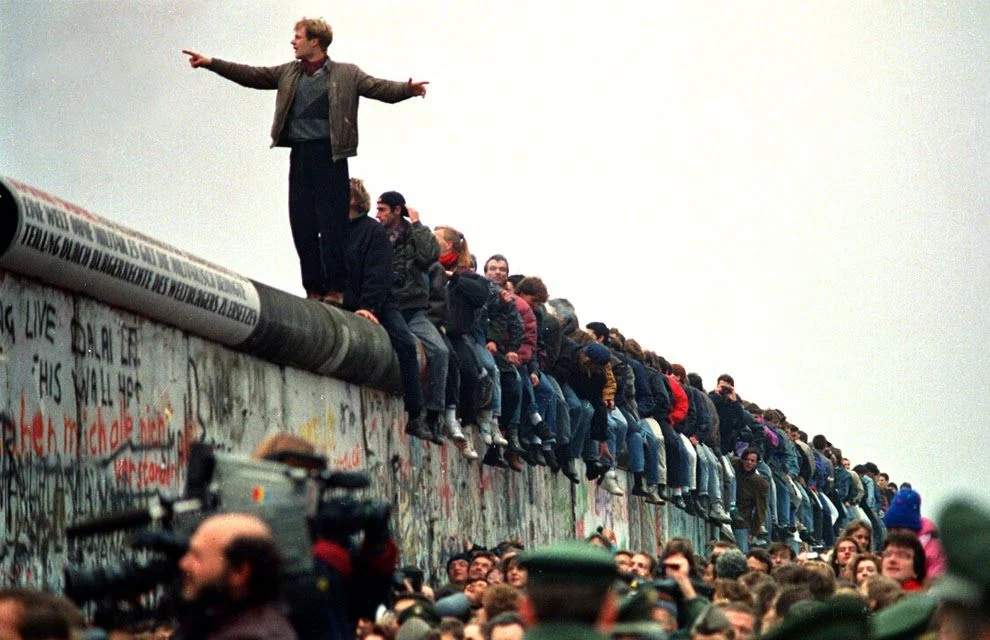 Berlineses sentados y de pie sobre el Muro de Berlín en noviembre de 1989, mientras periodistas filman desde abajo
