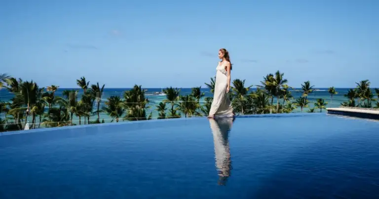 Mujer observando piscina de resort con palmeras y mar tropical