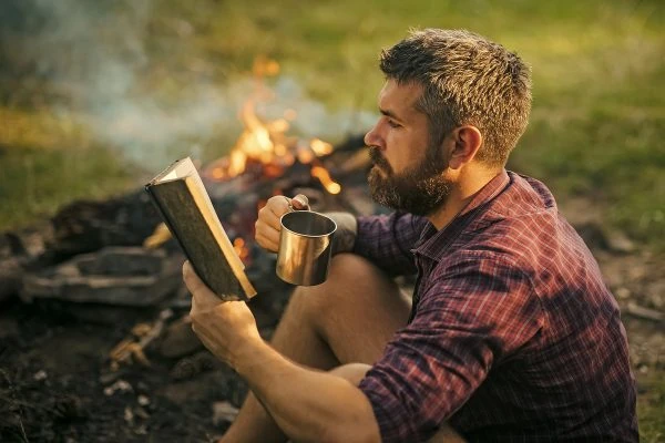 Hombre leyendo junto a fuego de campamento en la naturaleza