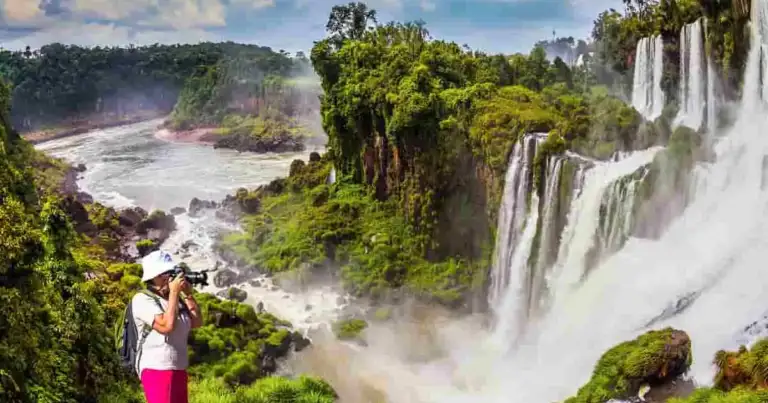 Turista con sombrero blanco fotografía varios saltos de las Cataratas del Iguazú, rodeados de selva, con el río Iguazú de fondo