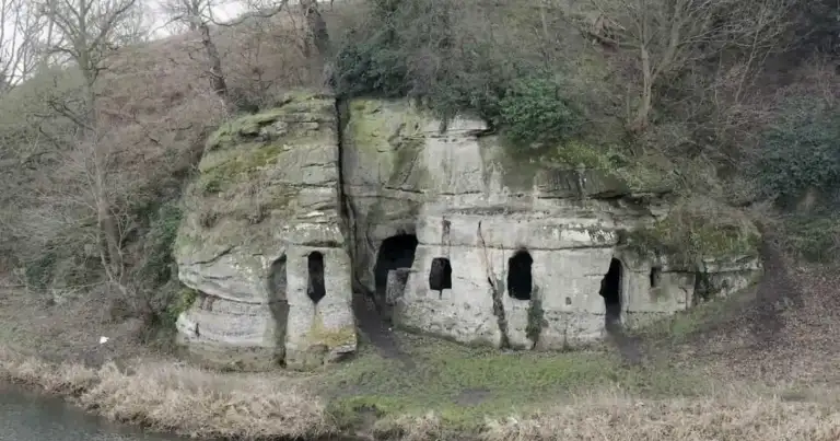 Formación rocosa con ventanas y puertas talladas, cubierta de musgo junto a un río