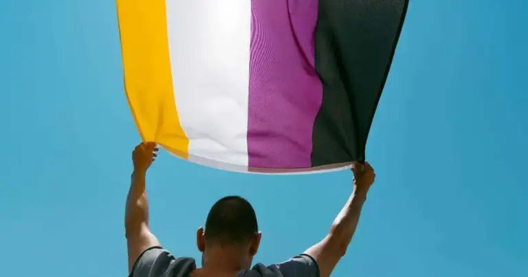 Persona sosteniendo bandera no binaria con franjas de múltiples colores contra cielo azul