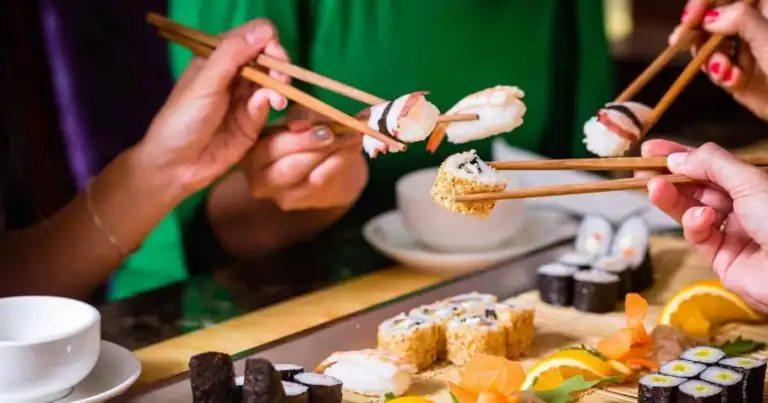 Manos sosteniendo palillos con sushi fresco sobre mesa con múltiples piezas de comida