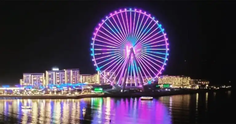 Noria iluminada en tonos púrpura y cian, reflejada en aguas oscuras de ciudad nocturna