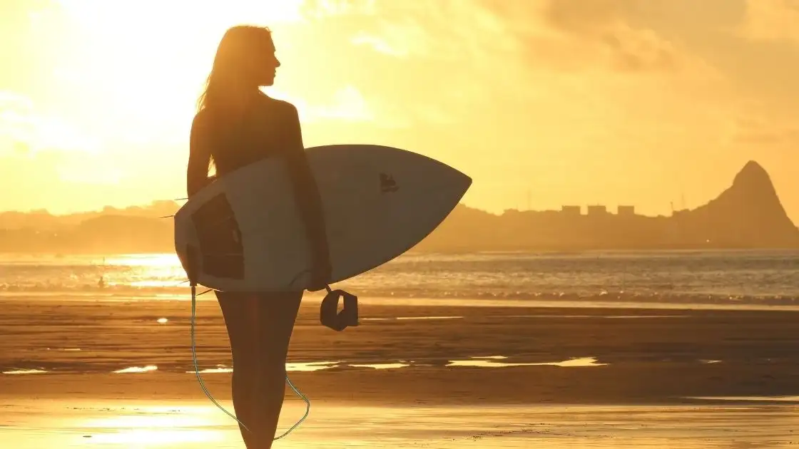 Surfista con tabla al atardecer en playa con silueta contra sol