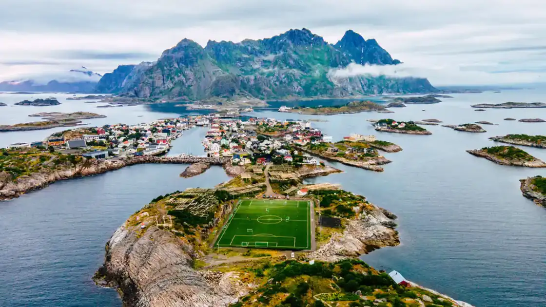 Isla rocosa con estadio de fútbol verde, pueblos y montañas al fondo sobre agua azul