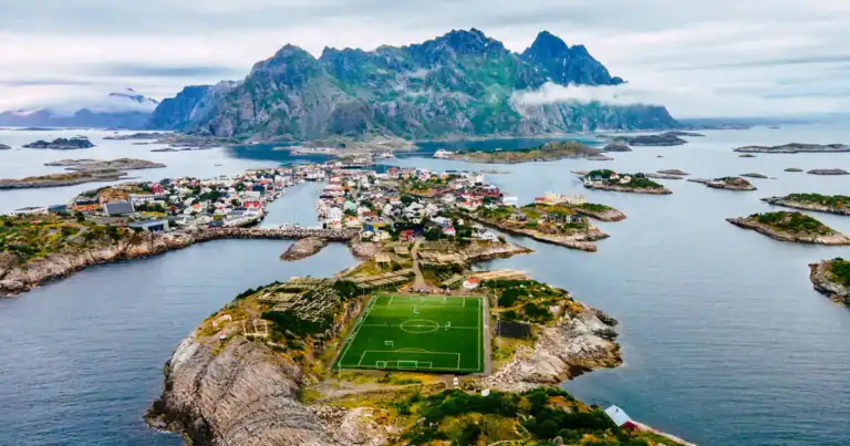 Isla rocosa con estadio de fútbol verde, pueblos y montañas al fondo sobre agua azul