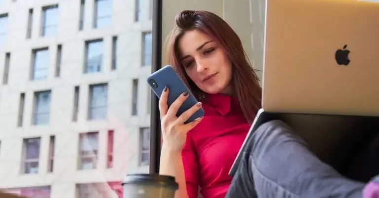 Mujer con teléfono azul y suéter rojo junto a laptop dorada