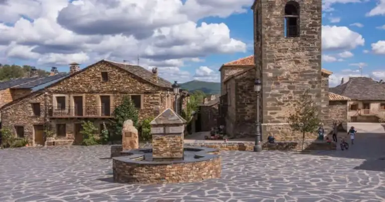 Plaza empedrada de un pueblo de casas de piedra, con una fuente y una torre de iglesia