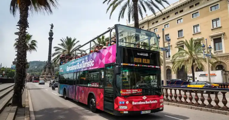 Autobús turístico de dos pisos recorriendo Barcelona, con la Sagrada Familia y La Pedrera al fondo, bajo el cielo azul.