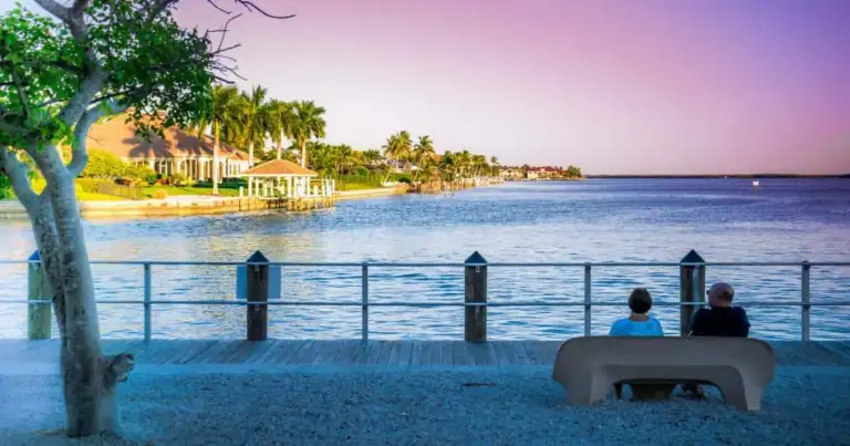 Playa costera con paseo marítimo, personas disfrutando el atardecer y casas al fondo