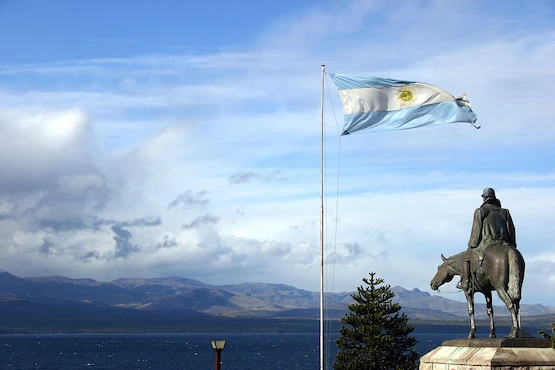 Bandera argentina ondeando junto a estatua ecuestre en Bariloche, con fondo de montañas y lago.