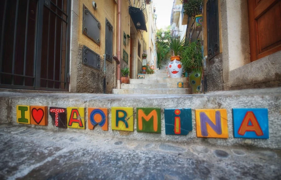 Calle de Taormina con letrero coloreado que dice "TAORMINA" en los bloques