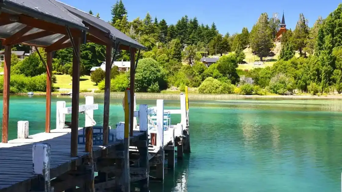 Muelle de madera sobre lago de aguas turquesas en Bariloche, rodeado de bosque y cabañas.
