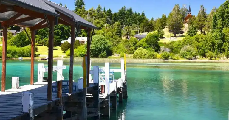 Muelle de madera sobre lago de aguas turquesas en Bariloche, rodeado de bosque y cabañas.