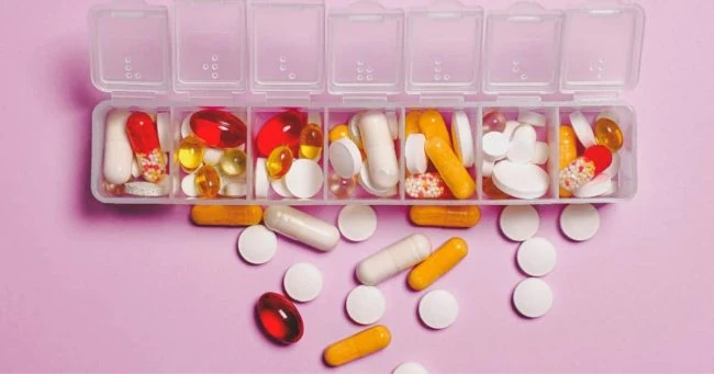 Organizador de medicamentos en compartimentos sobre fondo rosa con píldoras dispersas