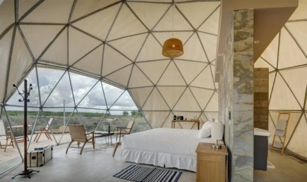 Domo geodésico con cama blanca y vistas a montañas, interior minimalista de glamping