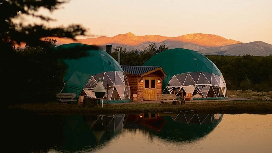 Domos verdes con edificio central reflejados en laguna al atardecer, montañas de fondo