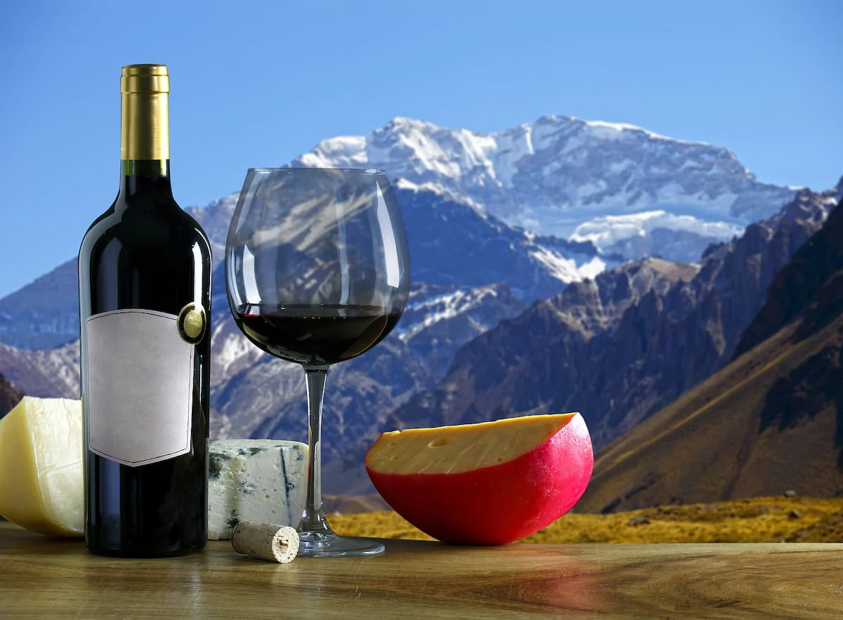 Botella de vino, copa de tinto y queso con montañas nevadas al fondo