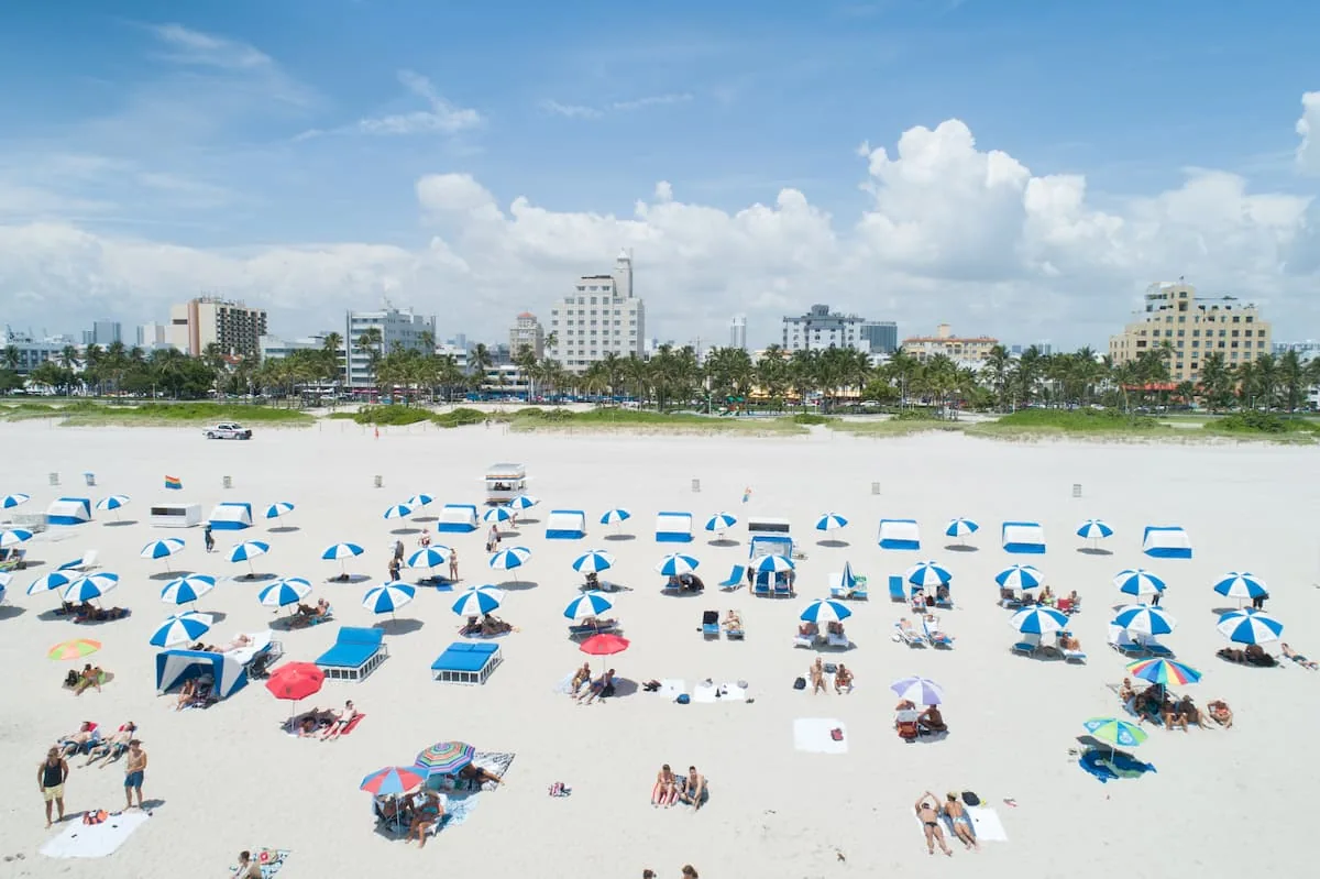 Playa llena de personas con sombrillas azules y edificios al fondo