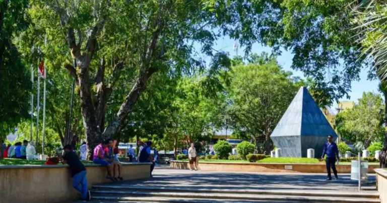 Parque con grandes árboles y estructura geométrica azul y blanca, personas caminando