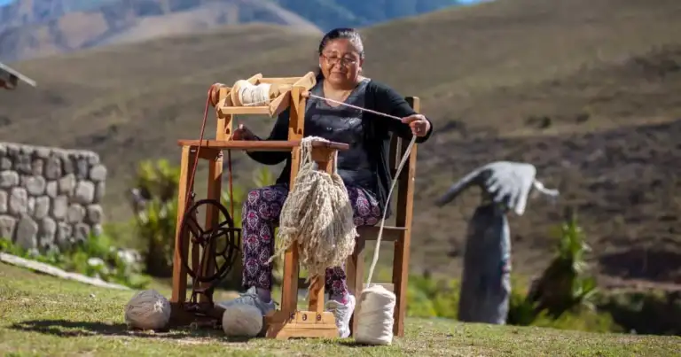 Mujer demostrando trabajo tradicional de textiles con niño y montañas al fondo