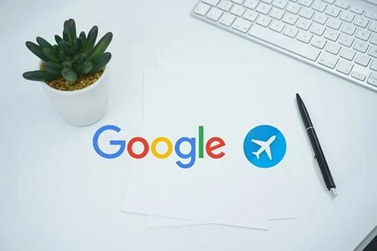 Escritorio blanco con logo Google, icono avión azul, planta y teclado