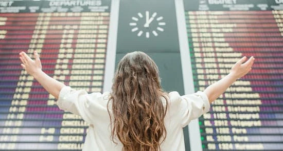 Persona de espaldas con brazos levantados mirando un tablero con horarios de vuelos en un aeropuerto