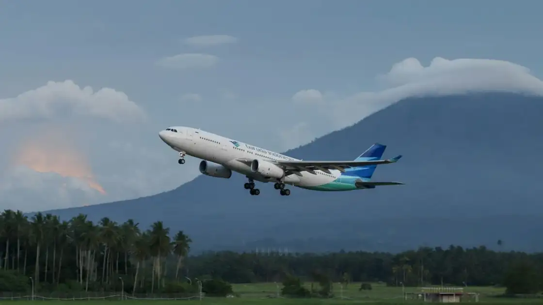 Avión de Garuda Indonesia despegando con montañas al fondo y palmeras en primer plano.