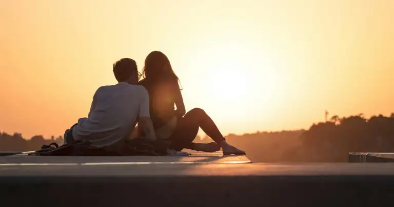 Pareja sentada al atardecer contra cielo naranja y agua tranquila