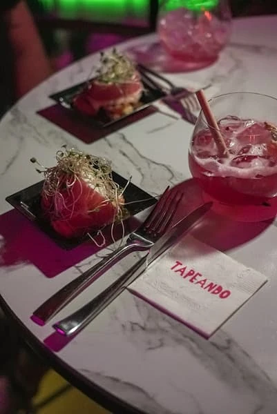 Tapas gourmet de jamón en platos negros con bebidas rosadas y mantel 'Tapeando'