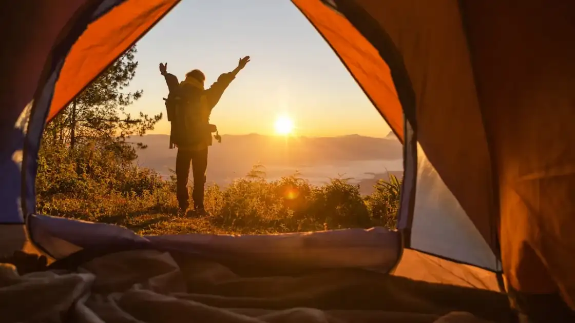 Excursionista se para frente a la tienda color naranja de campamento con la mochila mirando las montañas