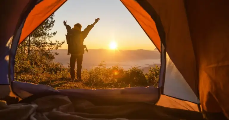 Excursionista se para frente a la tienda color naranja de campamento con la mochila mirando las montañas