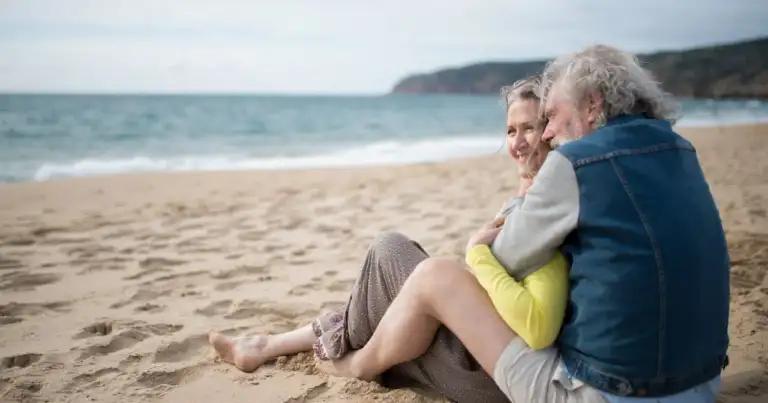 Pareja de adultos mayores abrazándose y sonriendo sentados en la playa.