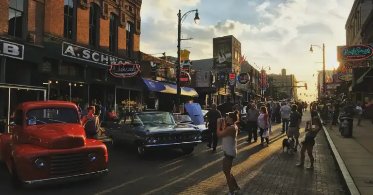 Personas caminando por Beale Street al atardecer en Memphis, rodeadas de autos clásicos y letreros de neón.