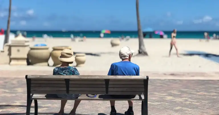 Pareja de turistas mayores sentados frente al mar en un día soleado, disfrutando de unas vacaciones relajadas en la playa.